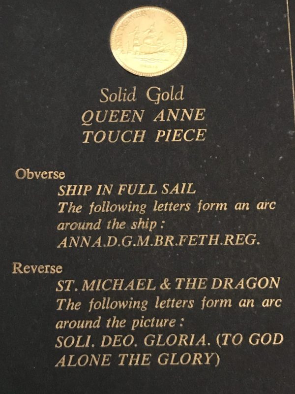 Display card with a gold coin at the top labeled “Solid Gold Queen Anne Touch Piece,” featuring descriptions of the coin’s obverse and reverse designs in gold lettering.