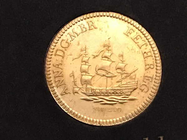 Close-up of a gold coin showing a detailed ship in full sail surrounded by the Latin inscription “ANNA D.G.M. BR. FET. H. REG.”