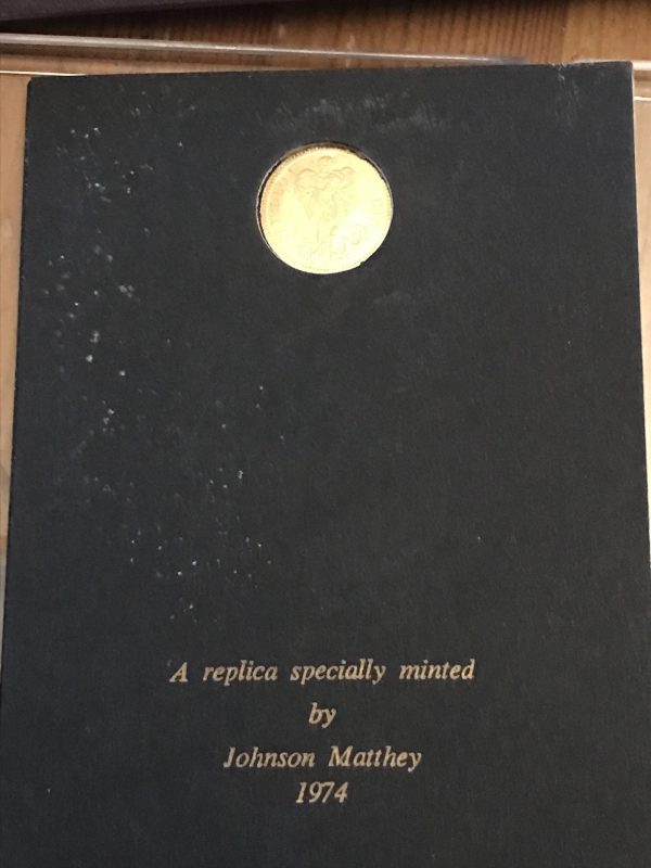 Black presentation card featuring a gold coin near the top and gold lettering below reading “A replica specially minted by Johnson Matthey 1974.”
