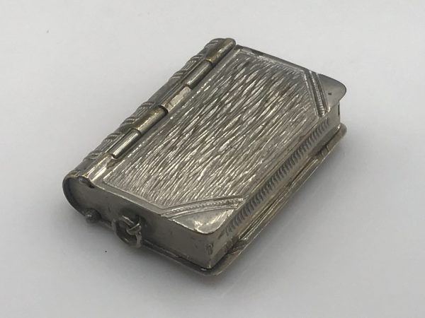 Angled close-up of an antique silver book-form vesta case, highlighting the textured surface and detailed hinge mechanism.