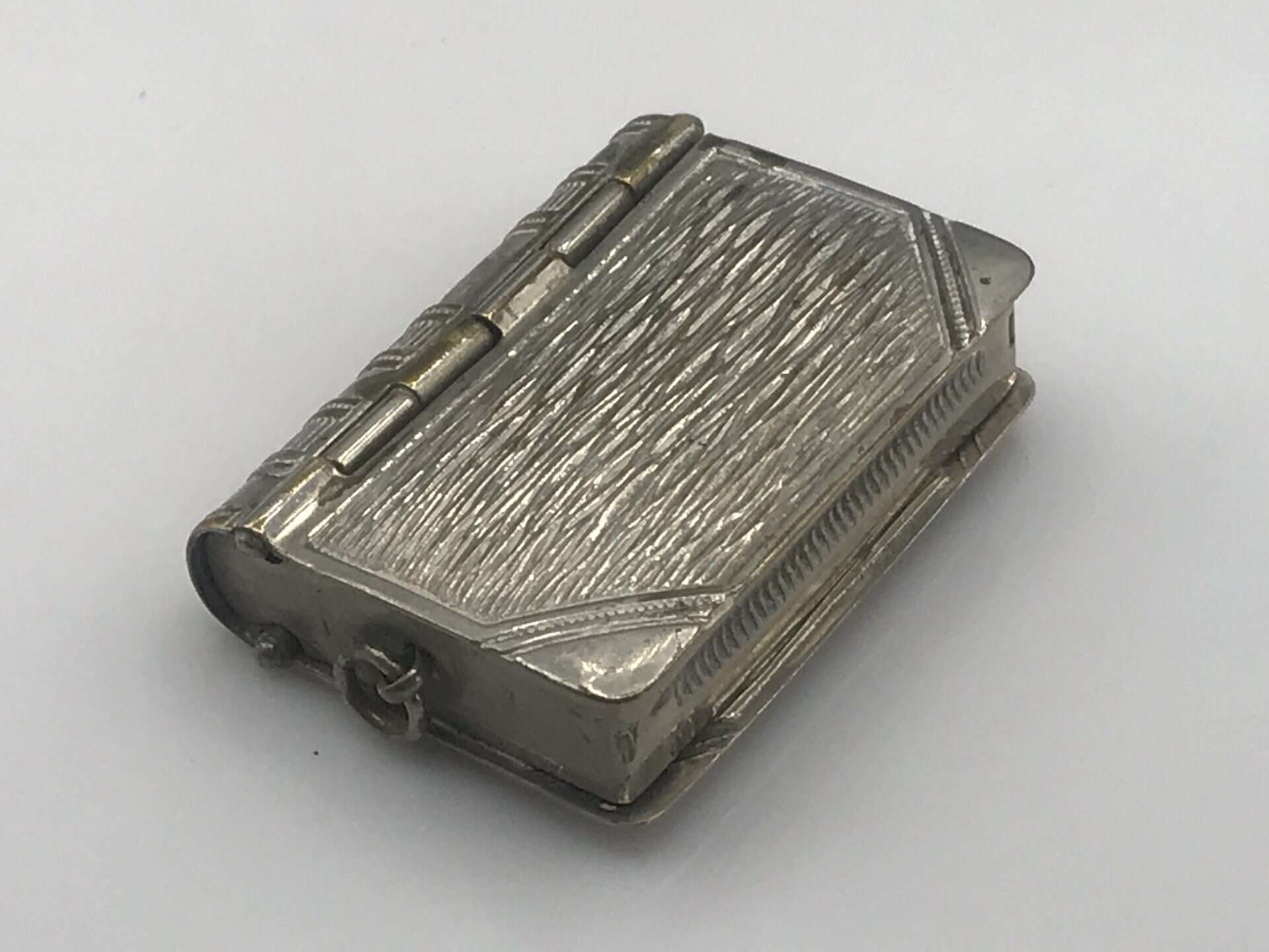 Angled close-up of an antique silver book-form vesta case, highlighting the textured surface and detailed hinge mechanism.