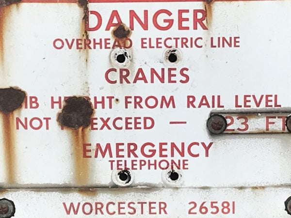 Close-up of antique enamel railway warning sign text in red on white enamel, showing safety instructions and Worcester emergency telephone number