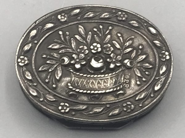 Close-up of antique 830 silver pill box lid showing repoussé floral basket design and silver 830 hallmark