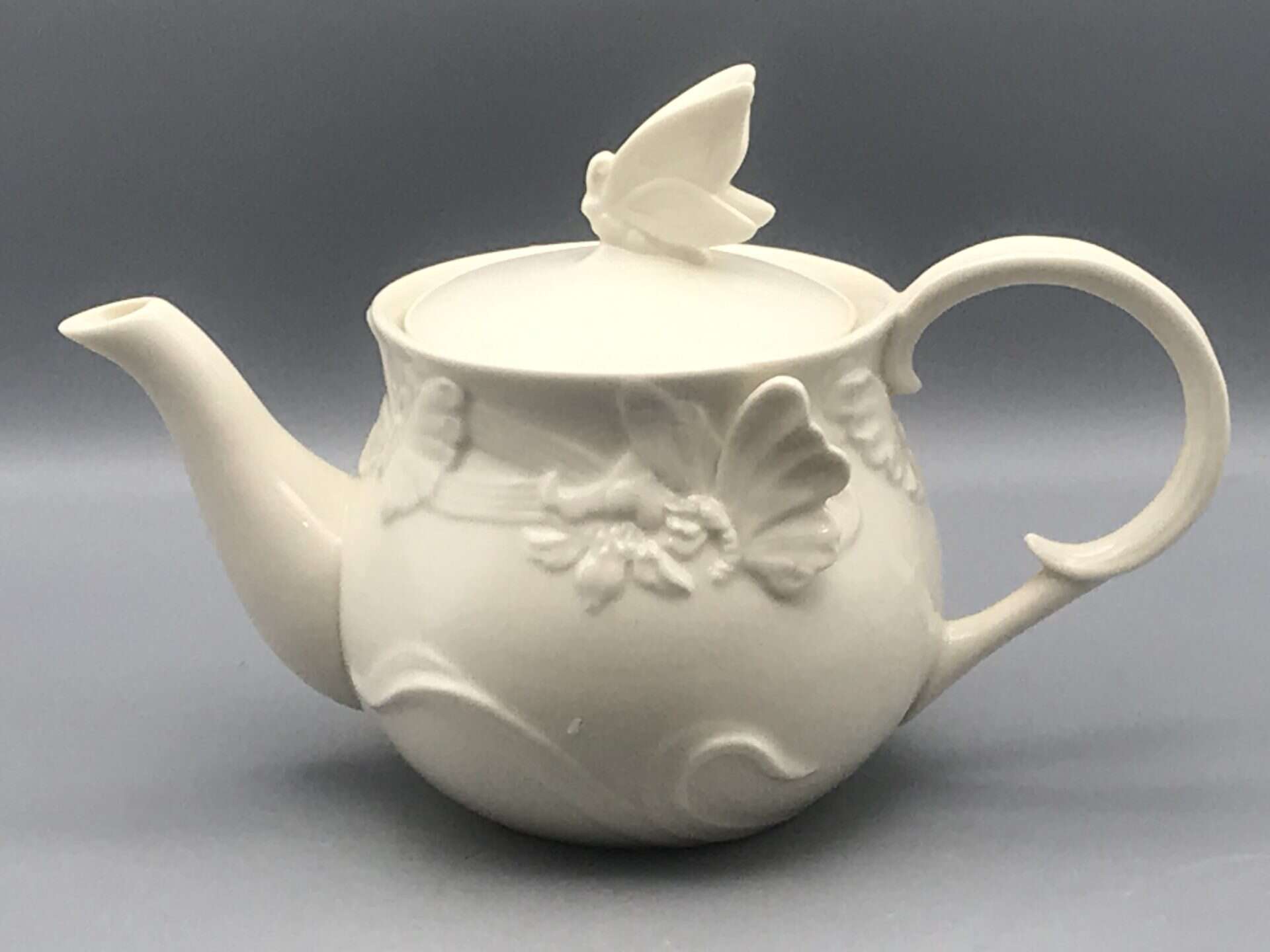 Vintage Regal ceramic teapot with sculpted butterfly design in ivory glaze, featuring butterfly finial and curved spout, approximately 9.25 inches long and 5.75 inches tall.