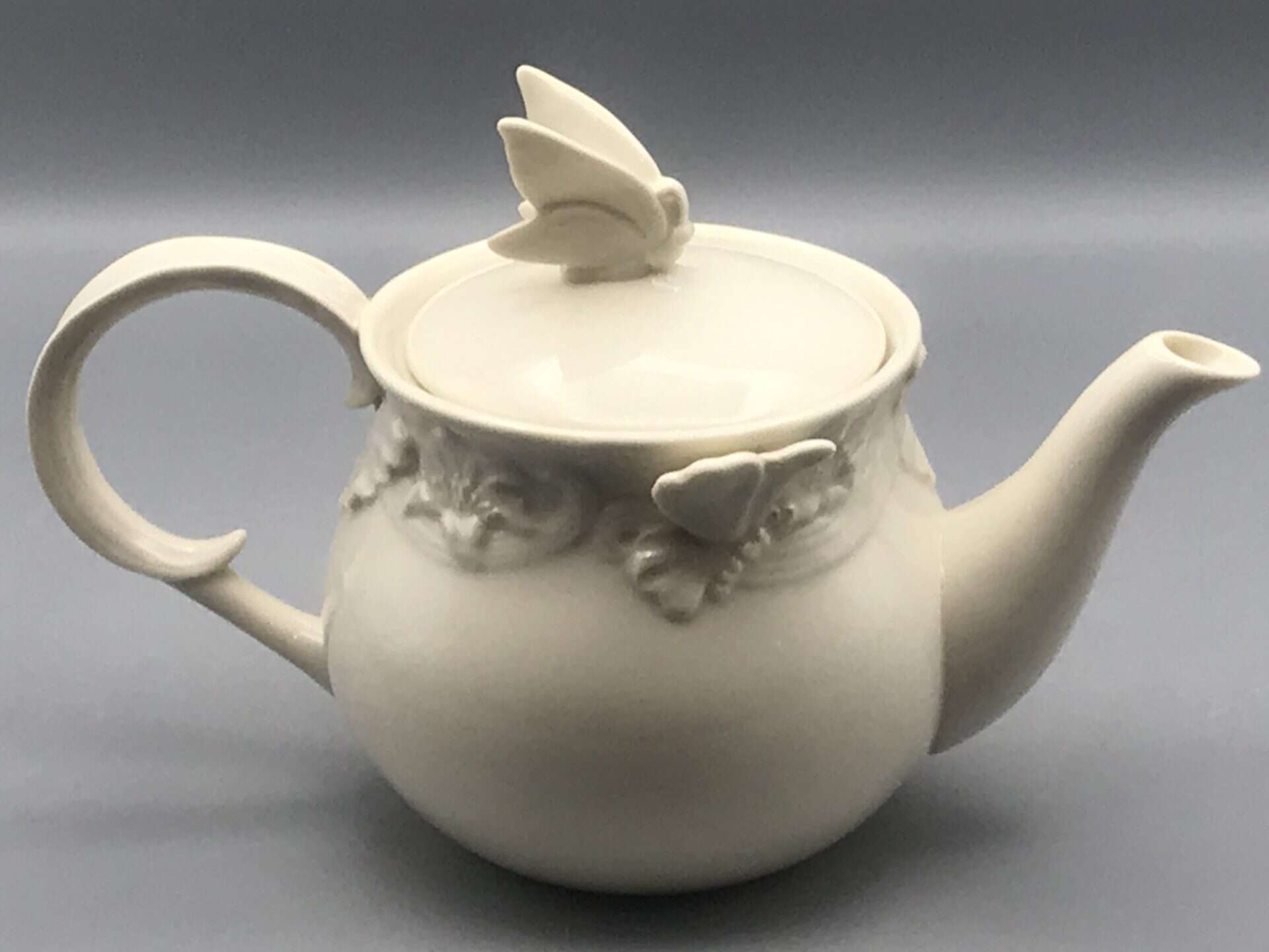 Side view of vintage Regal ceramic teapot with ivory glaze, showing butterfly relief motifs and butterfly finial on the lid.