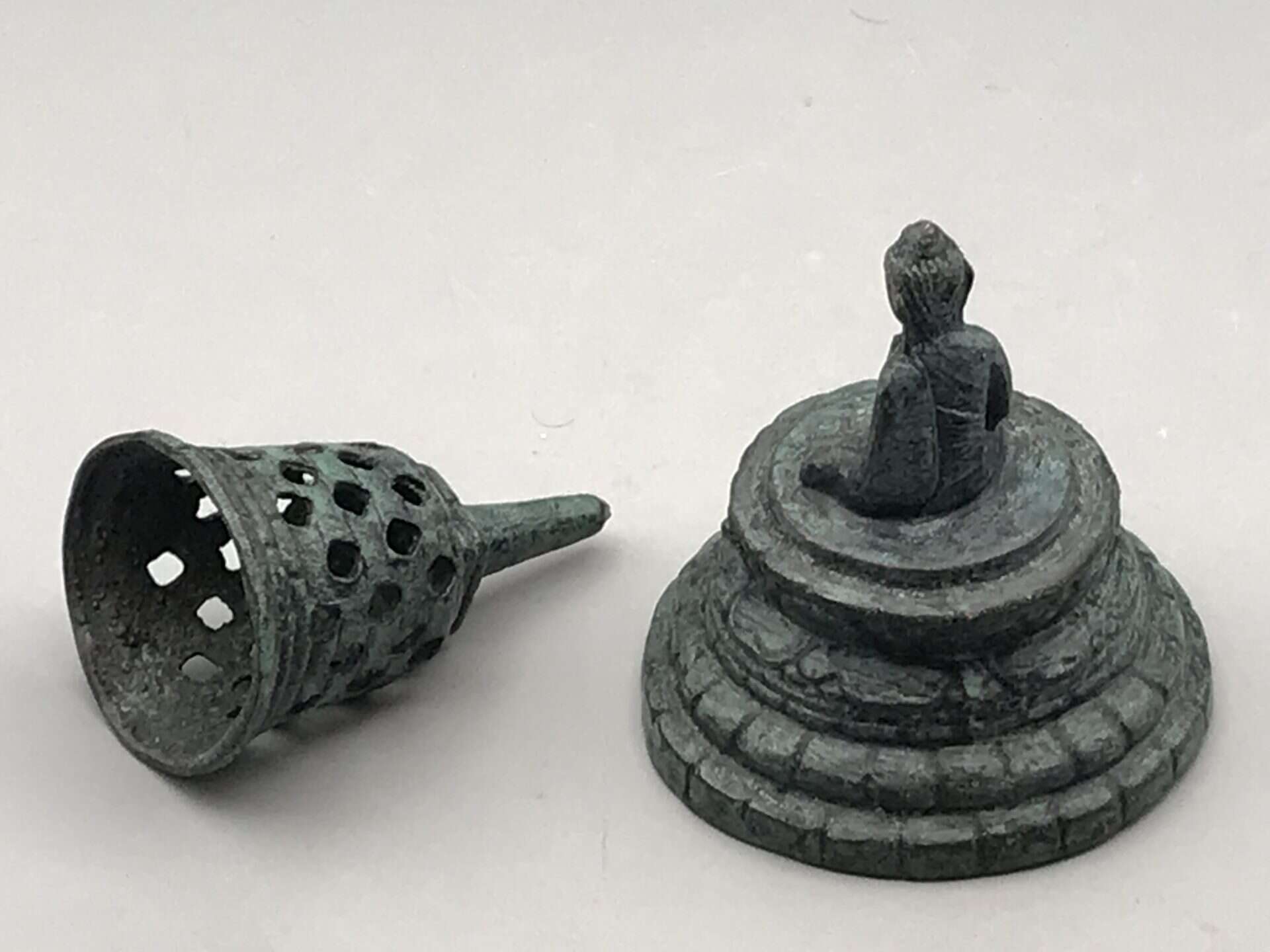 Bronzed base metal stupa bell shown disassembled, with the pierced cover lying beside the base that supports a seated Buddha figure.