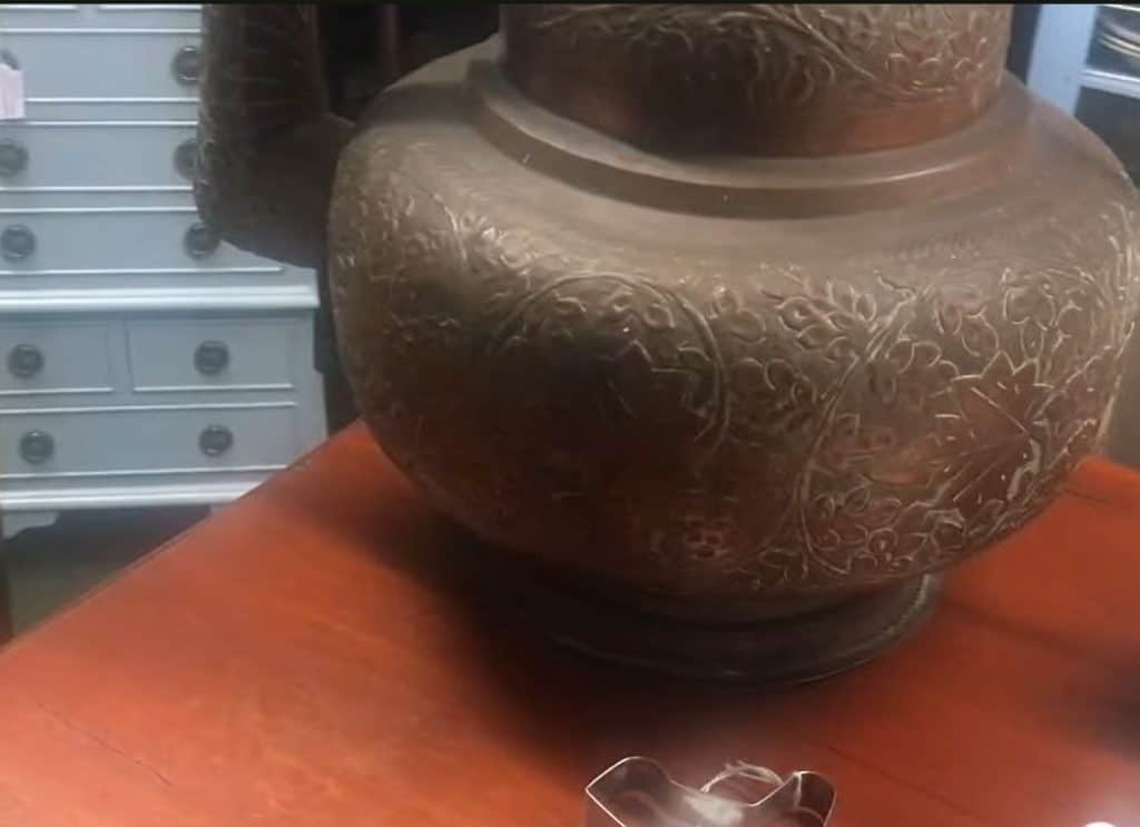 Bottom half of a monumental late 19th-century Ottoman copper ewer resting on a wooden table inside an antique centre, showing the hand-raised bulbous body with repoussé floral decoration and original aged patina at the time of discovery