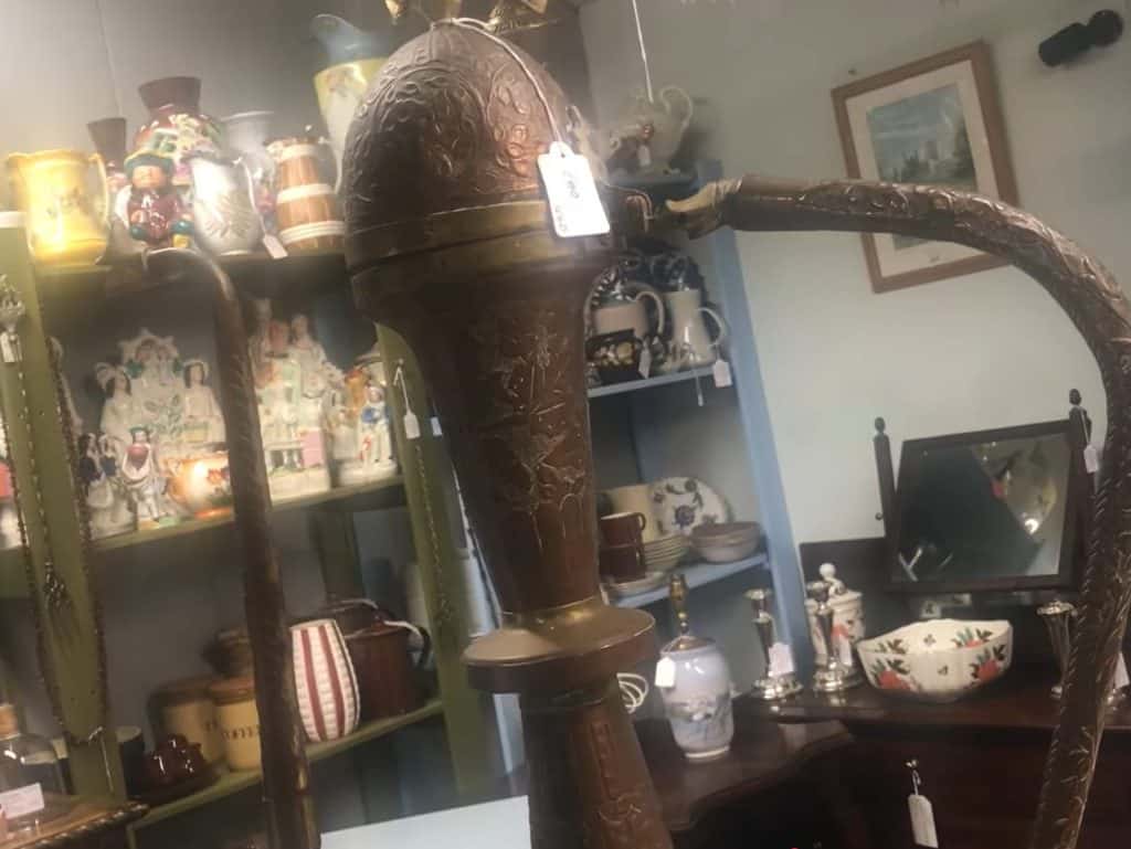 Upper section of a monumental late 19th-century Ottoman copper ewer standing prominently in the centre of an antique centre, showing the tall baluster neck, domed lid with finial, swan-head spout, curved handle, and original price tag at the time of discovery